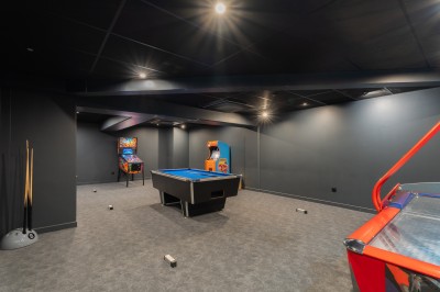 Salle lumineuse avec canapés, fauteuils, table basse, étagère de livres et écran mural, sur un sol en moquette grise.