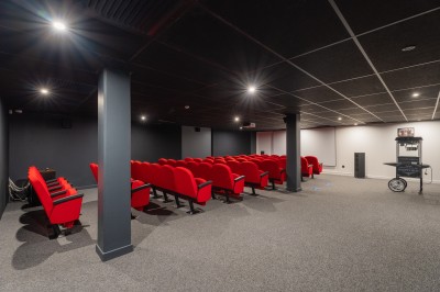 Salle lumineuse avec canapés, fauteuils, table basse, étagère de livres et écran mural, sur un sol en moquette grise.