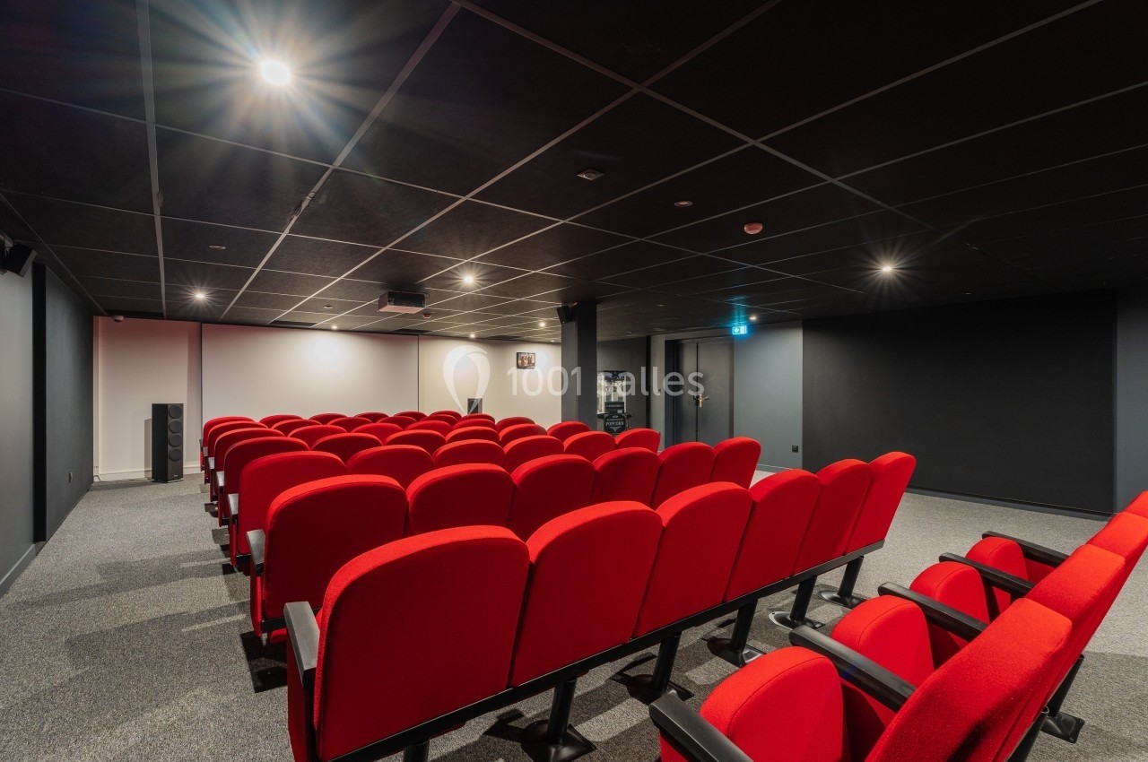 Salle de projection avec rangées de fauteuils rouges alignés, éclairage tamisé et écran blanc à l'avant.