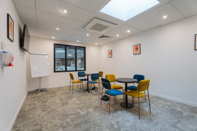 Salle lumineuse avec canapés, fauteuils, table basse, étagère de livres et écran mural, sur un sol en moquette grise.