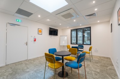 Salle lumineuse avec canapés, fauteuils, table basse, étagère de livres et écran mural, sur un sol en moquette grise.