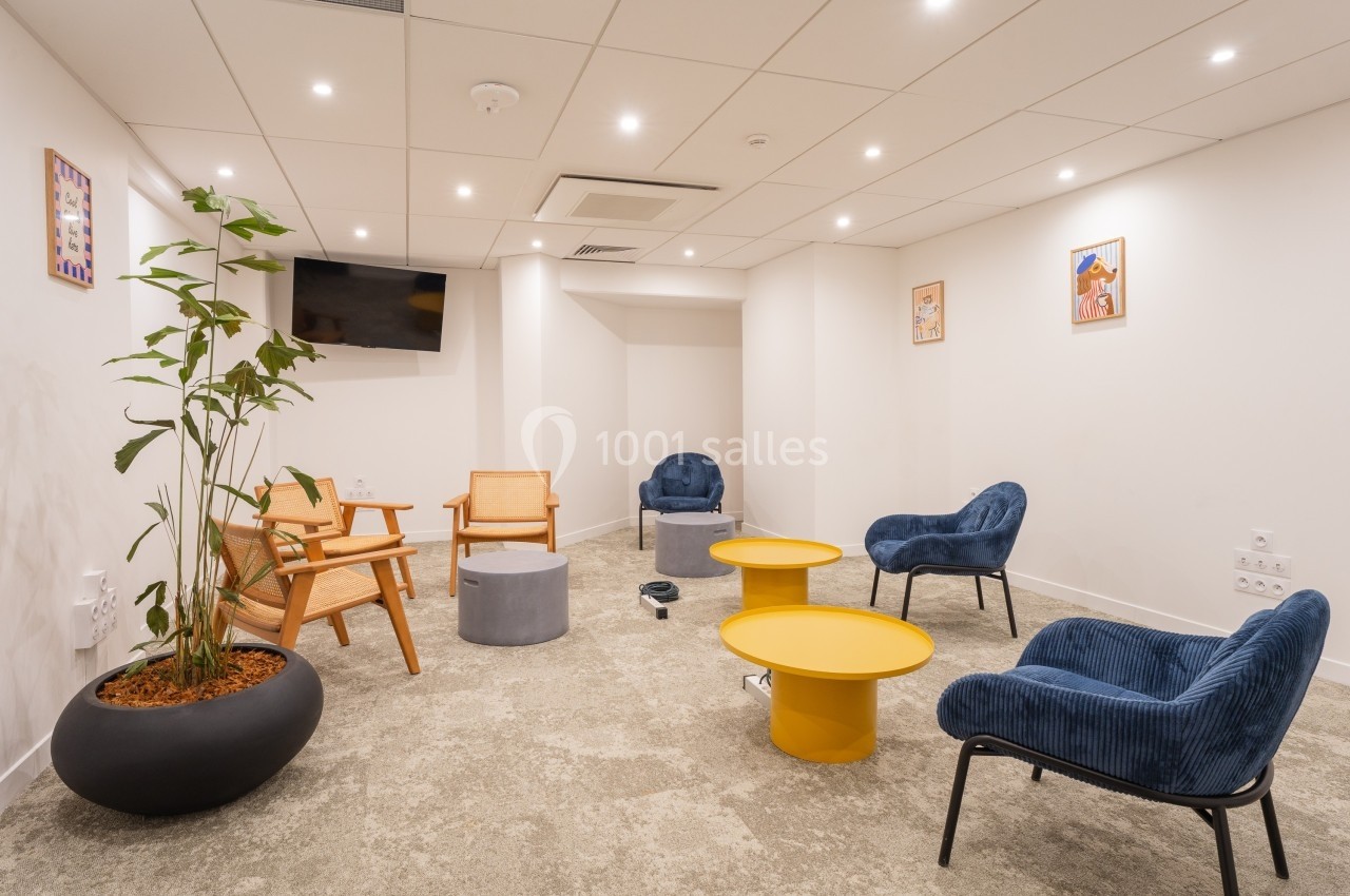Espace de détente avec fauteuils bleus, chaises en bois, tables basses jaunes et plantes dans un intérieur lumineux.