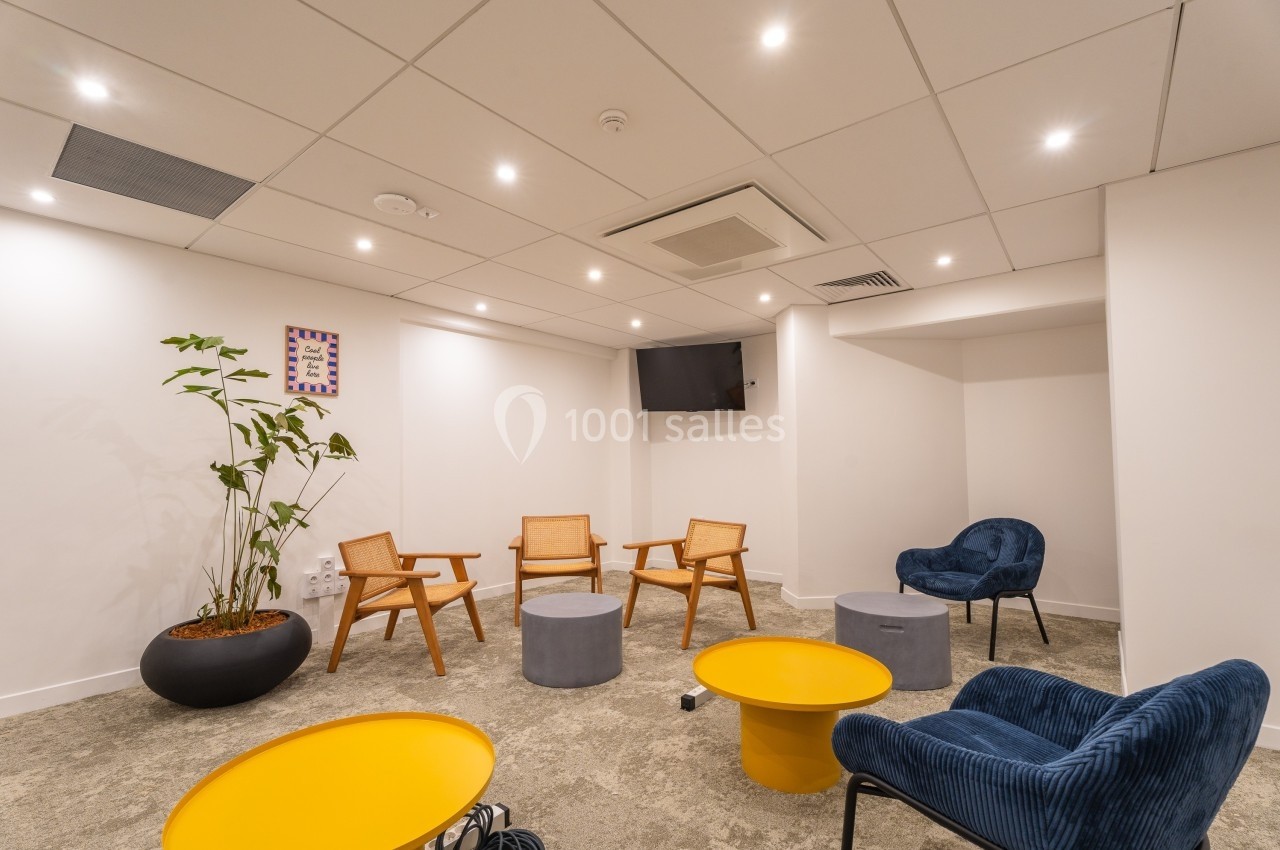 Salle lumineuse avec fauteuils en bois et en velours, tables basses jaunes et grises, et une plante en pot.