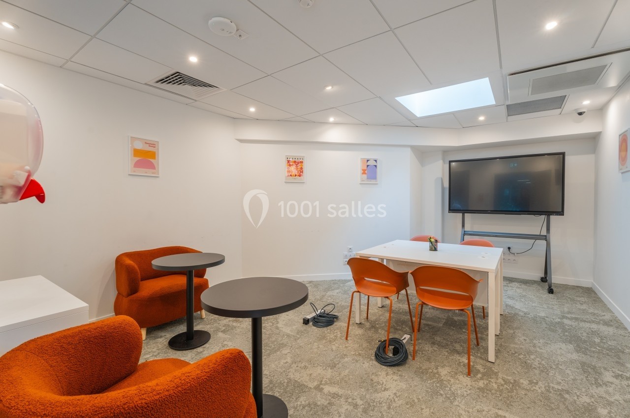 Salle de réunion moderne avec table blanche, chaises orange, fauteuils, écran mural et décor minimaliste.
