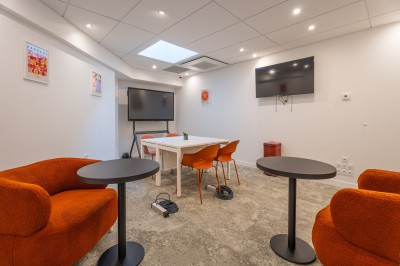 Salle lumineuse avec canapés, fauteuils, table basse, étagère de livres et écran mural, sur un sol en moquette grise.