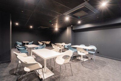 Salle lumineuse avec canapés, fauteuils, table basse, étagère de livres et écran mural, sur un sol en moquette grise.