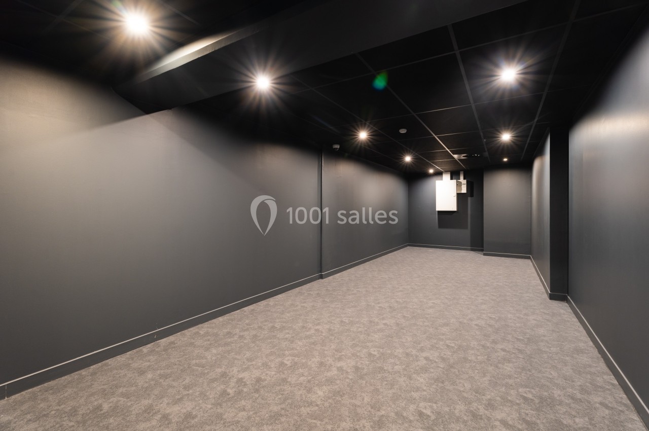 Salle vide avec murs et plafond noirs, sol gris, éclairée par des spots encastrés au plafond.