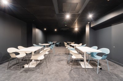Salle lumineuse avec canapés, fauteuils, table basse, étagère de livres et écran mural, sur un sol en moquette grise.
