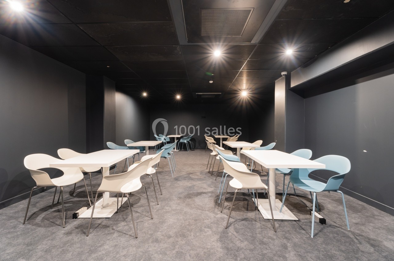 Salle moderne avec des murs sombres, éclairage au plafond, tables blanches et chaises design beige et bleues.