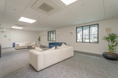 Salle lumineuse avec canapés, fauteuils, table basse, étagère de livres et écran mural, sur un sol en moquette grise.