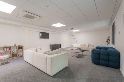 Salle lumineuse avec canapés, fauteuils, table basse, étagère de livres et écran mural, sur un sol en moquette grise.