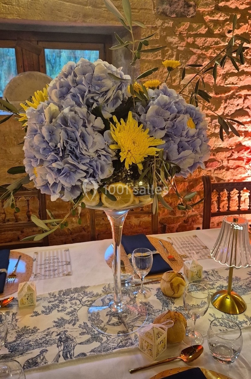 Centre de table floral avec hortensias bleus, fleurs jaunes et feuillage, posé sur un verre haut dans une salle en pierre.