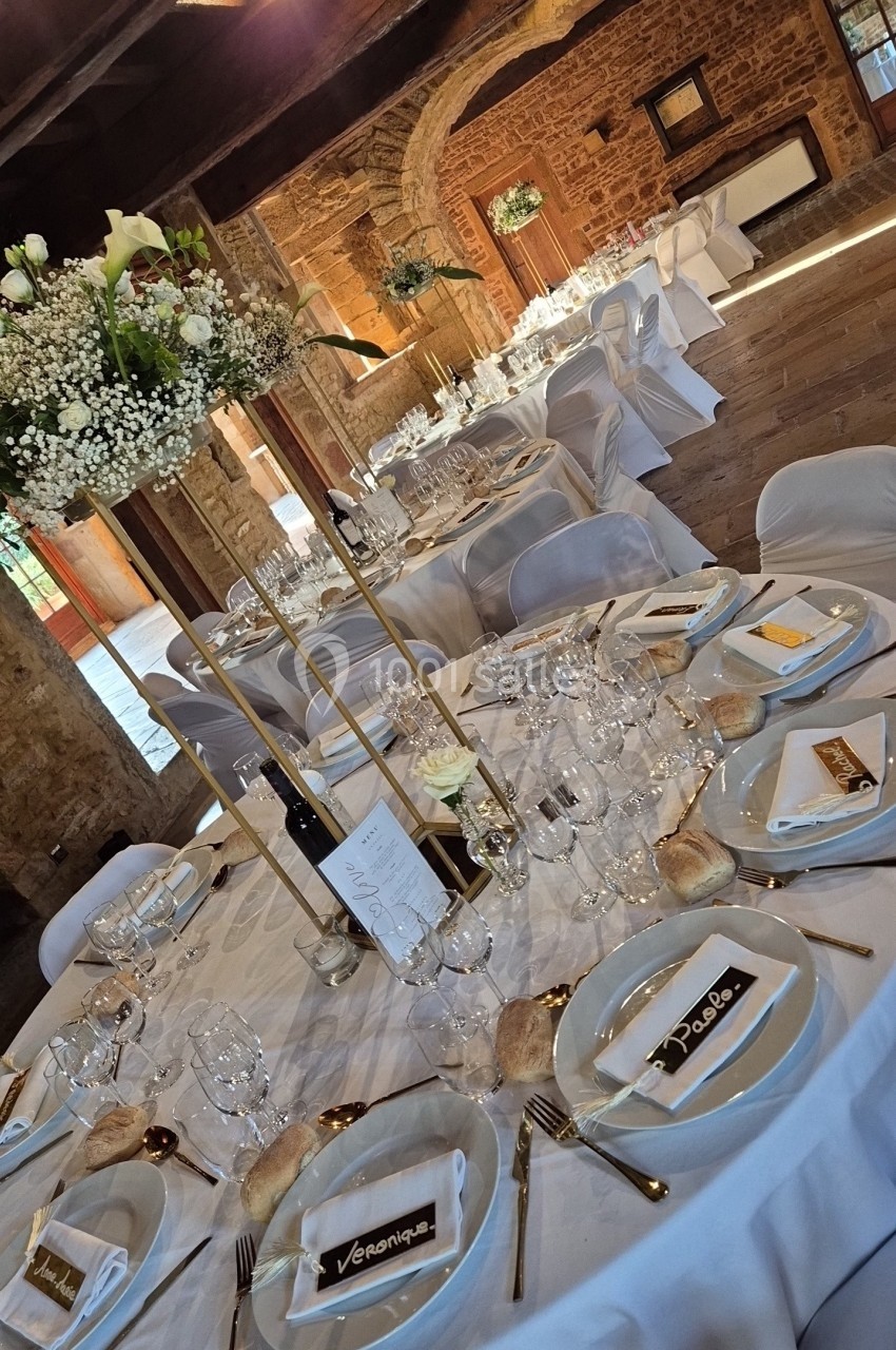 Salle de réception rustique avec tables rondes dressées, nappes blanches, décor floral et menus personnalisés.