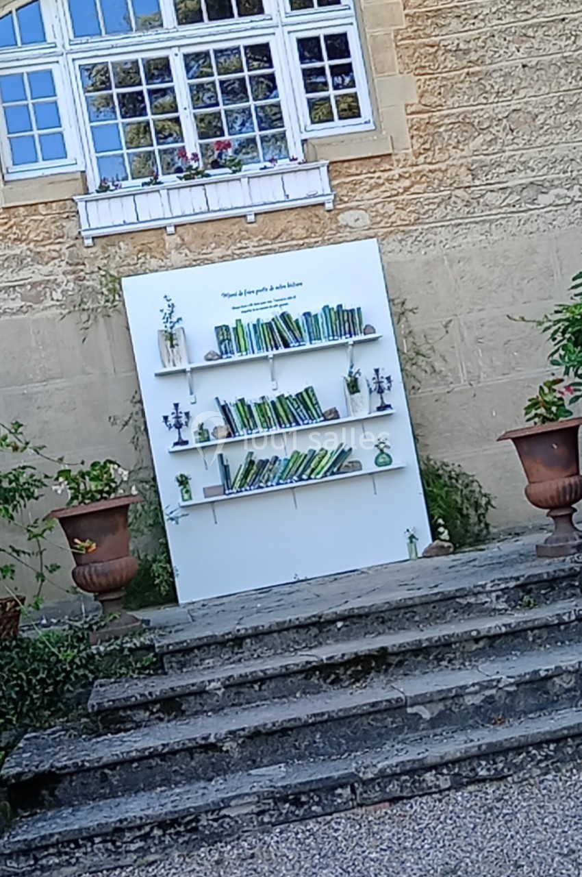 Étagère blanche avec des livres et des plantes, installée en extérieur devant un mur en pierre et une fenêtre.