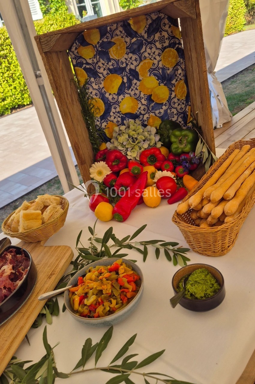 Table décorée avec des légumes frais, fruits, pain, tapenade et charcuterie, sur fond de carreaux colorés.