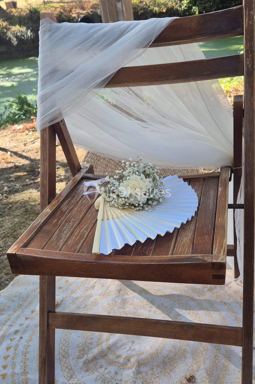 Chaise en bois décorée de tissu blanc, avec un éventail et un bouquet de fleurs blanches posés sur l'assise.