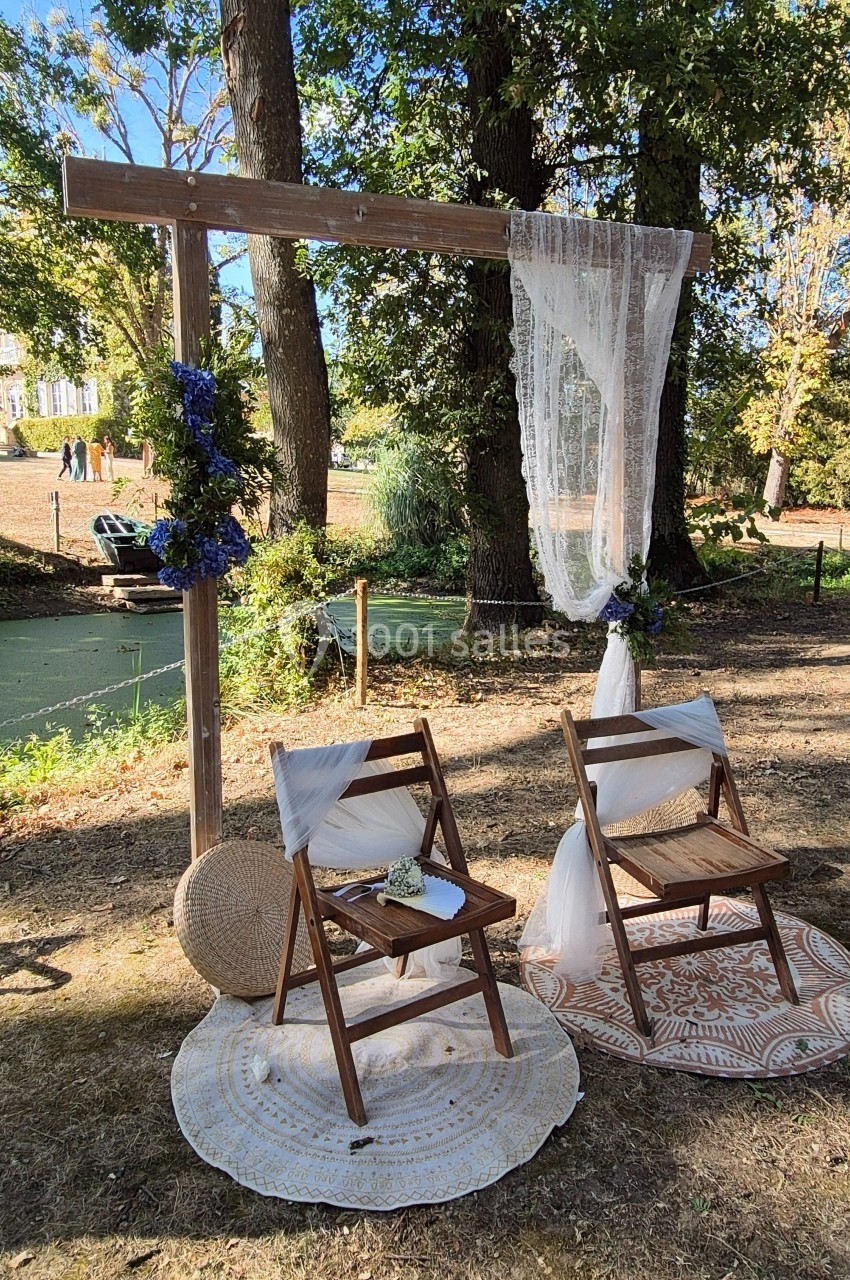 Deux chaises en bois décorées sous une arche ornée de fleurs et de voilages, installées en extérieur près d'arbres.
