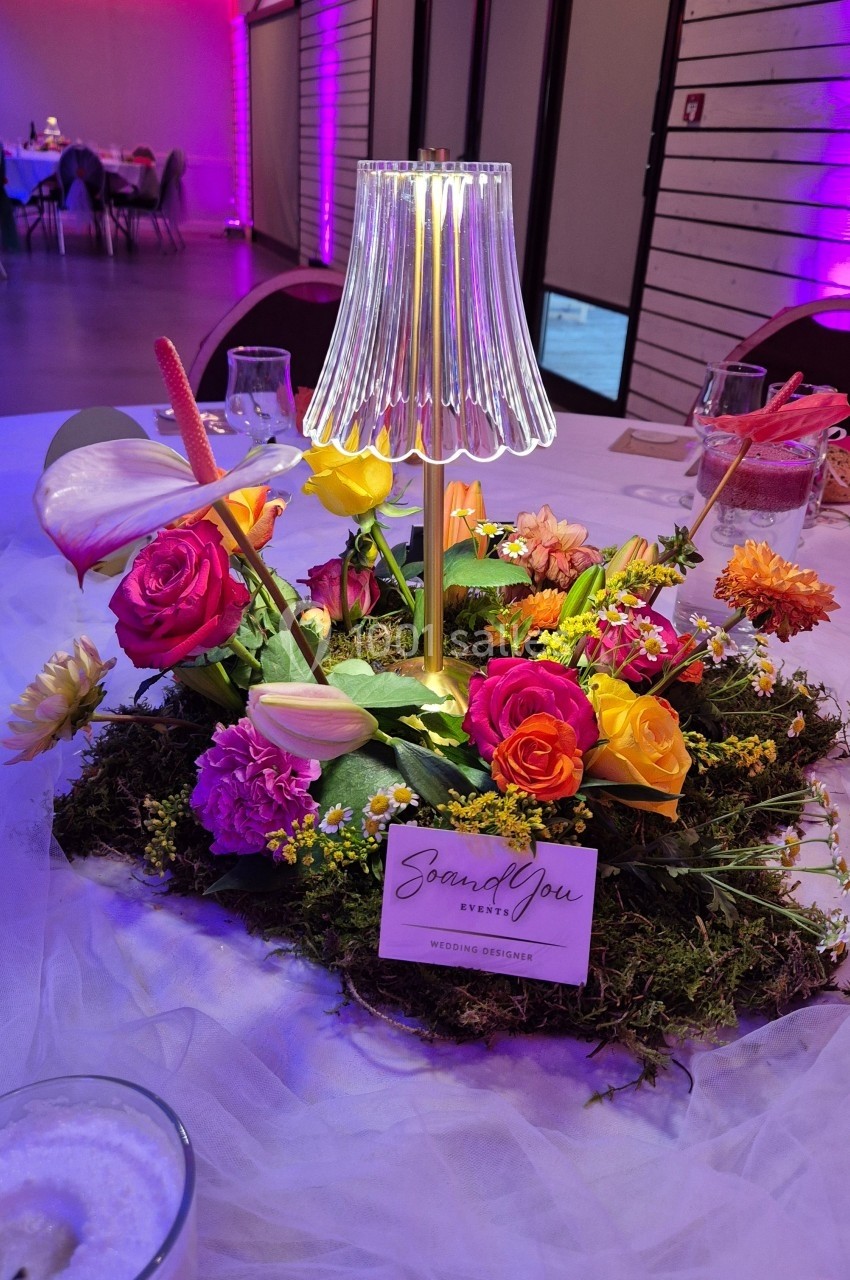 Centre de table floral coloré avec une lampe à abat-jour transparent, posé sur une nappe blanche dans une salle éclairée.