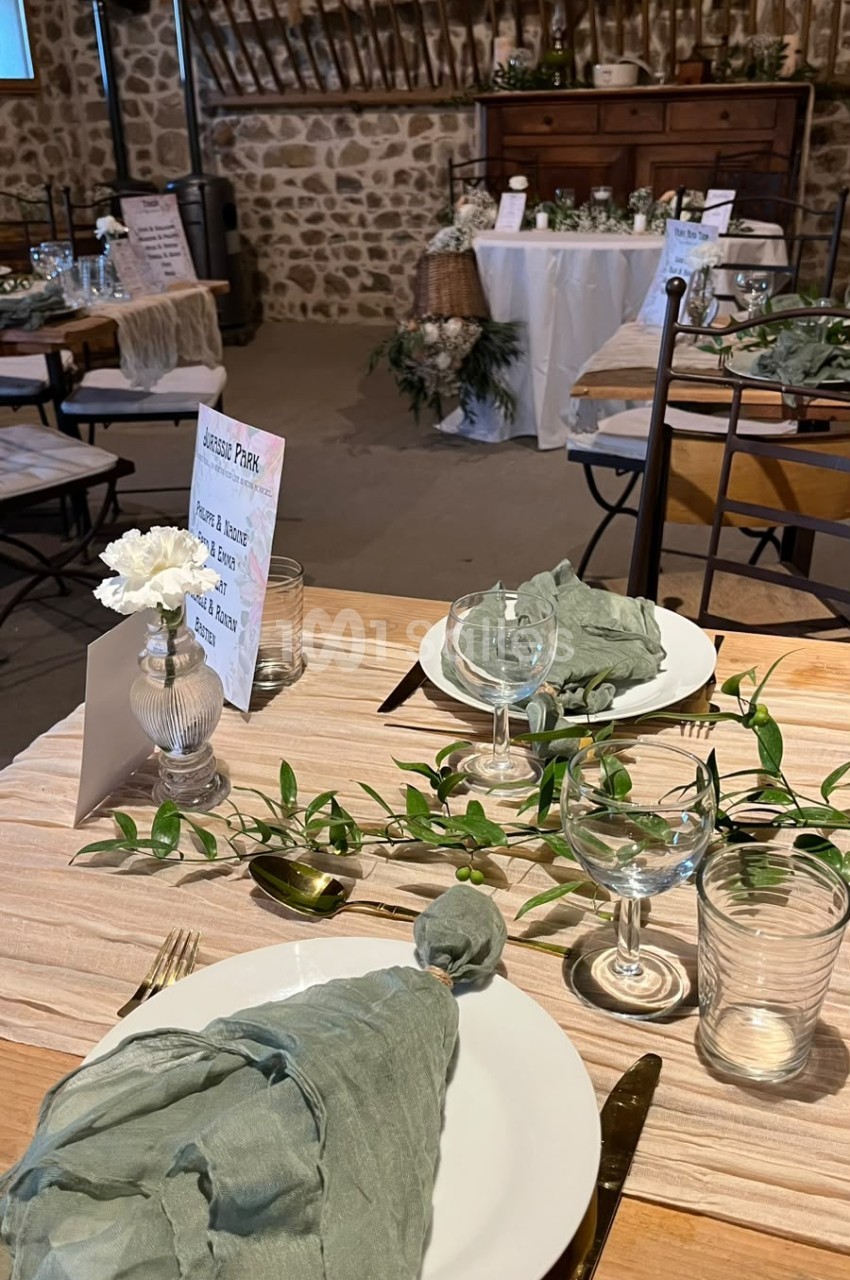 Table dressée en bois clair avec chemin de table en lin, serviette vert sauge, verres, guirlande de feuillage et vase à œillet blanc, dans une salle aux murs en pierre.