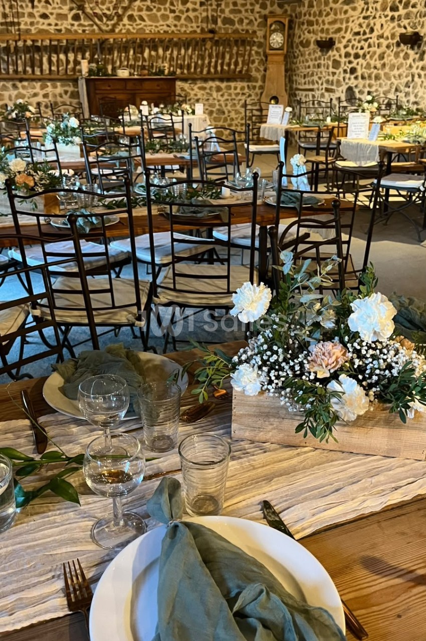 Table de réception dressée en bois avec chemin de table en lin, assiettes blanches, serviettes vert sauge et centre floral de roses blanches et eucalyptus dans une caisse en bois, dans une salle aux murs de pierre.