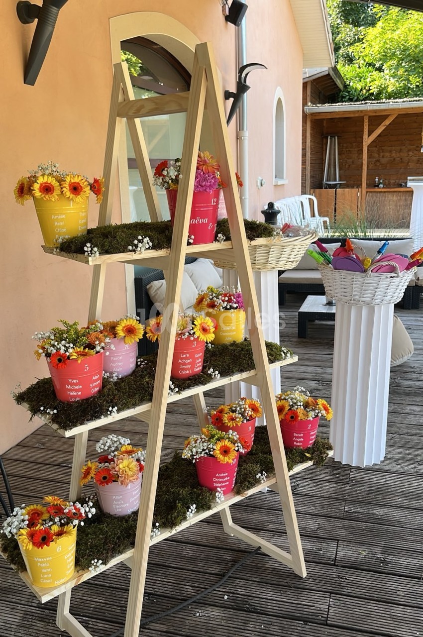 Escalier en bois à quatre niveaux garni de seaux colorés fleuris de gerberas jaunes, orange et roses, posé en terrasse extérieure.