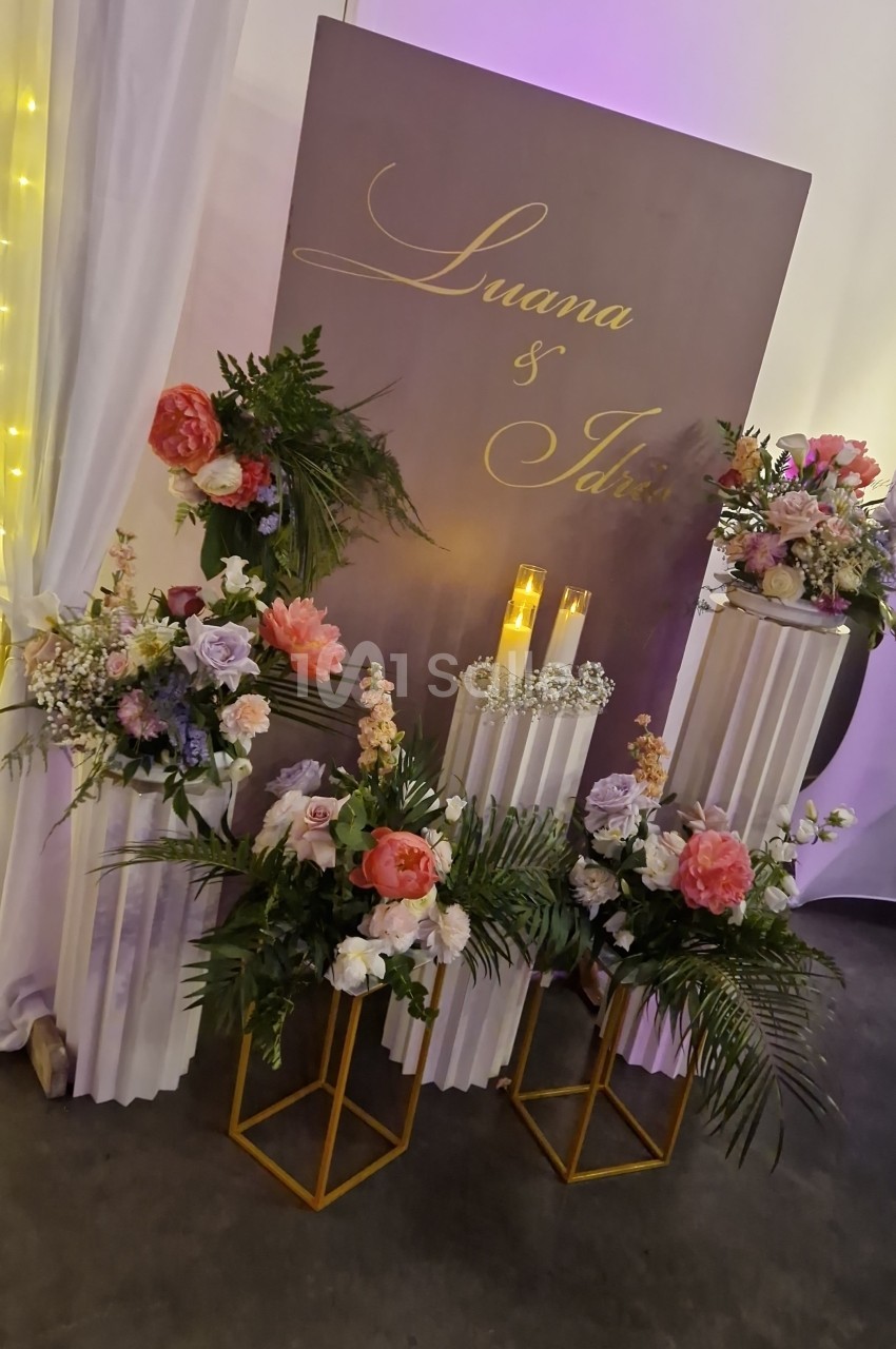 Panneau taupe à lettrage doré entouré de bouquets corail, roses et lilas sur piédestaux blancs et supports dorés.