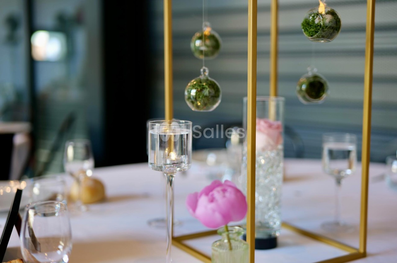 Table dressée de décoration dorée : cadres en métal doré, globes de verre avec mousse verte suspendus, pivoine rose et bougies sur une nappe blanche.