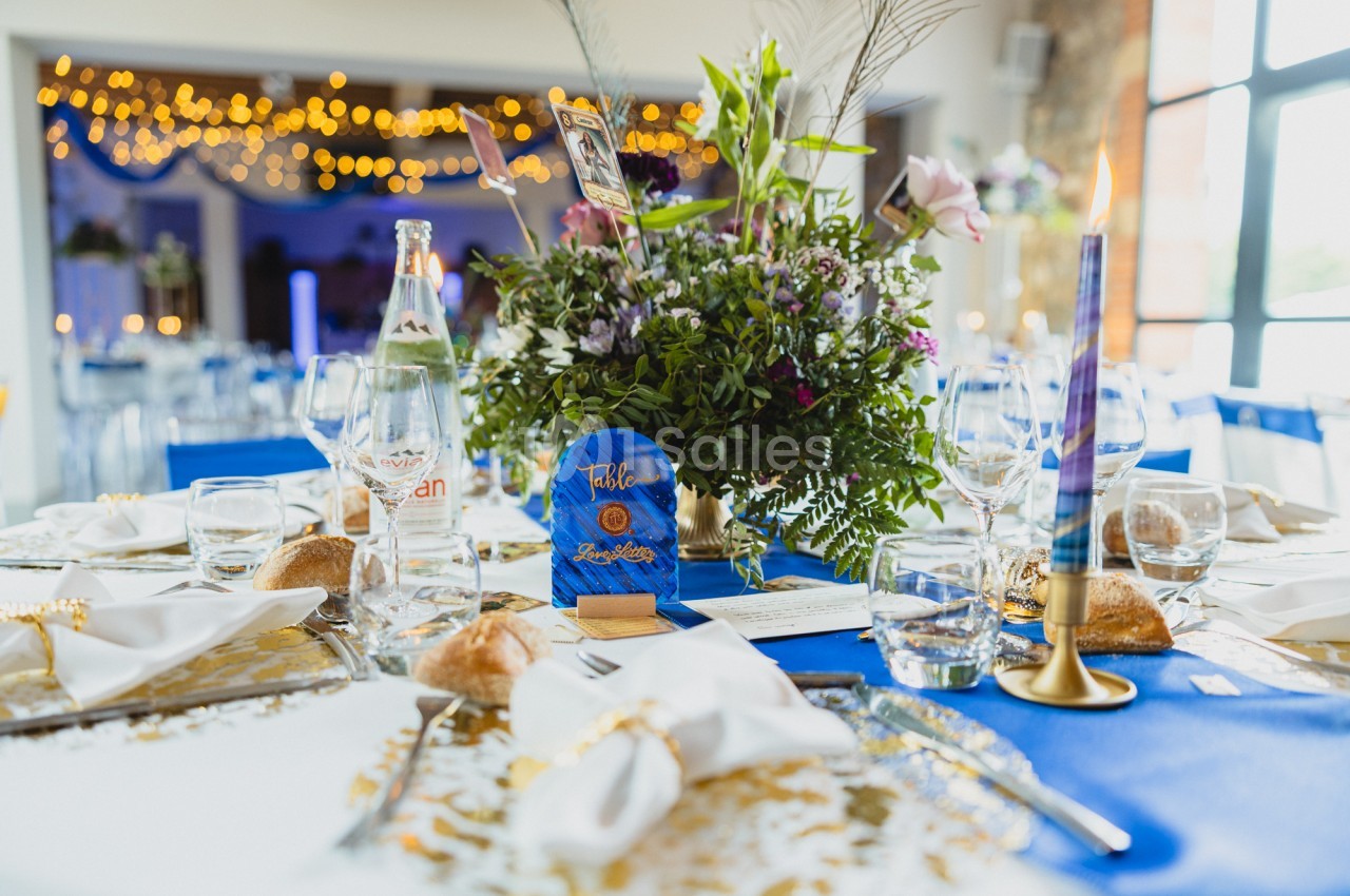 Table de réception à nappe bleue, bouquet de fleurs et feuillage au centre, bougie haute et serviettes blanches.