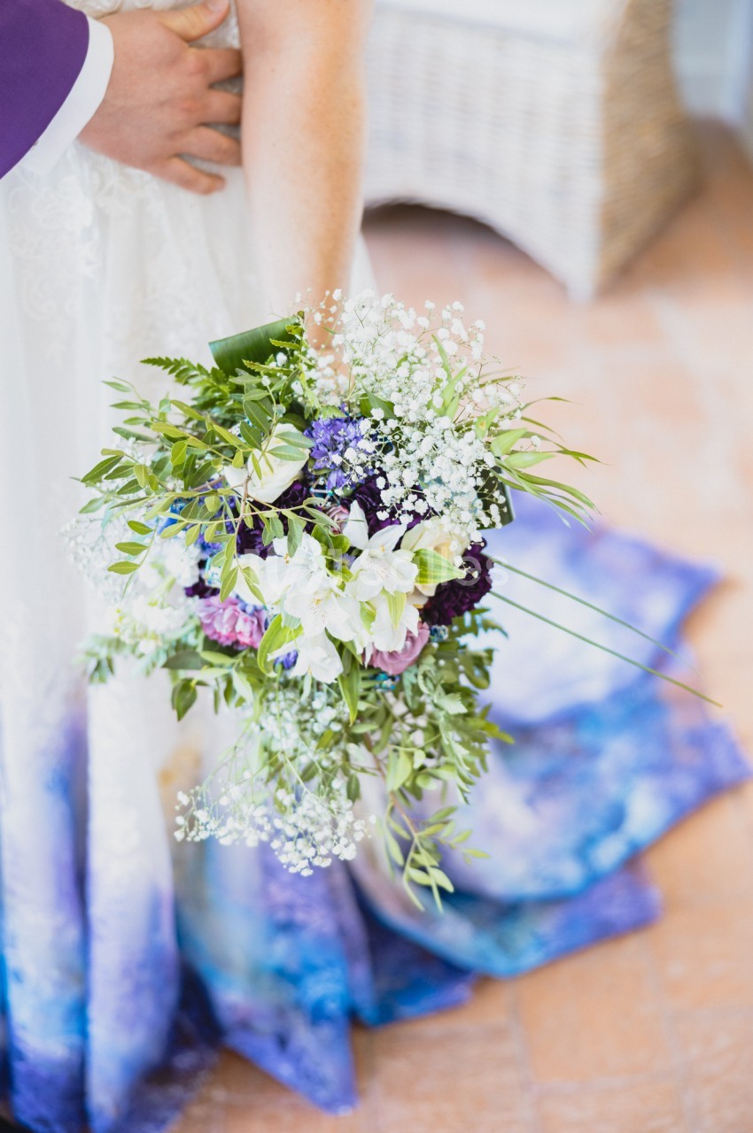 Bouquet de mariée cascadant aux fleurs blanches, violettes et roses, entouré de feuillage vert, tenu contre une robe blanche à traîne bleue.
