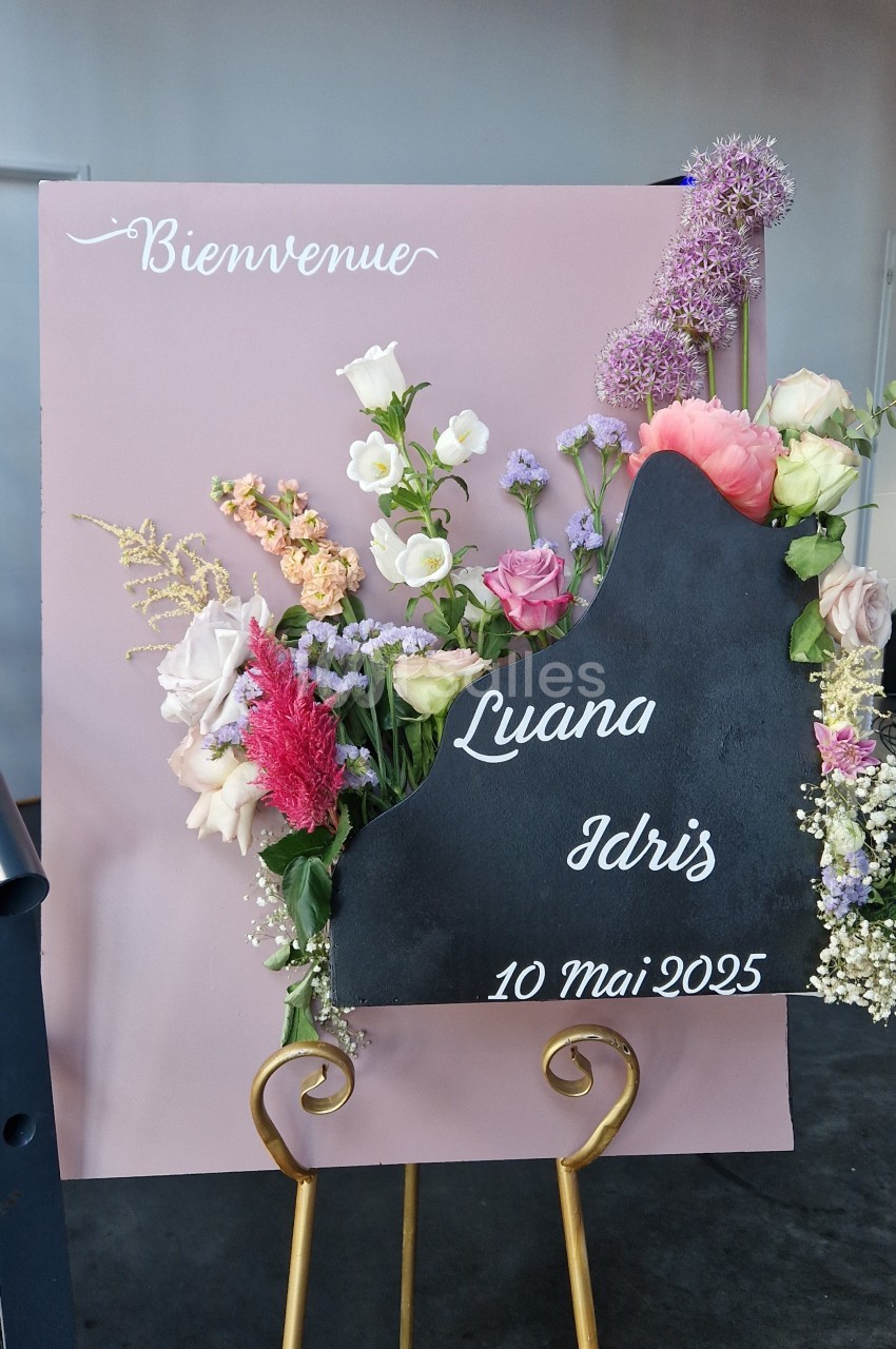 Panneau d'accueil rose poudré orné de fleurs fraîches (roses, pivoines, alliums) et d'une ardoise noire sur chevalet doré.