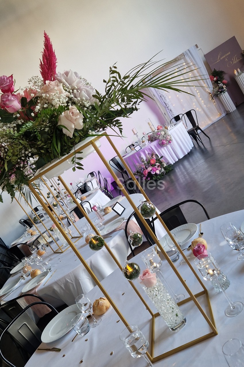 Table de réception dressée de nappes blanches, centres de table hauts en structure dorée ornés de fleurs roses, de boules végétales et de vases en cristal, avec en arrière-plan une table d'honneur éclairée de guirlandes lumineuses. Hmm, that's too long.