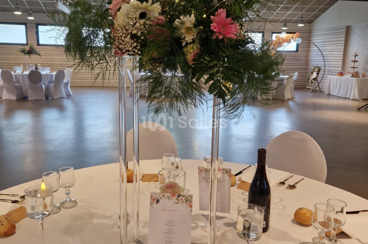 Table ronde dressée de nappes blanches avec un grand bouquet floral rose et vert sur colonnes en verre, dans une salle lumineuse aux chaises housses blanches.