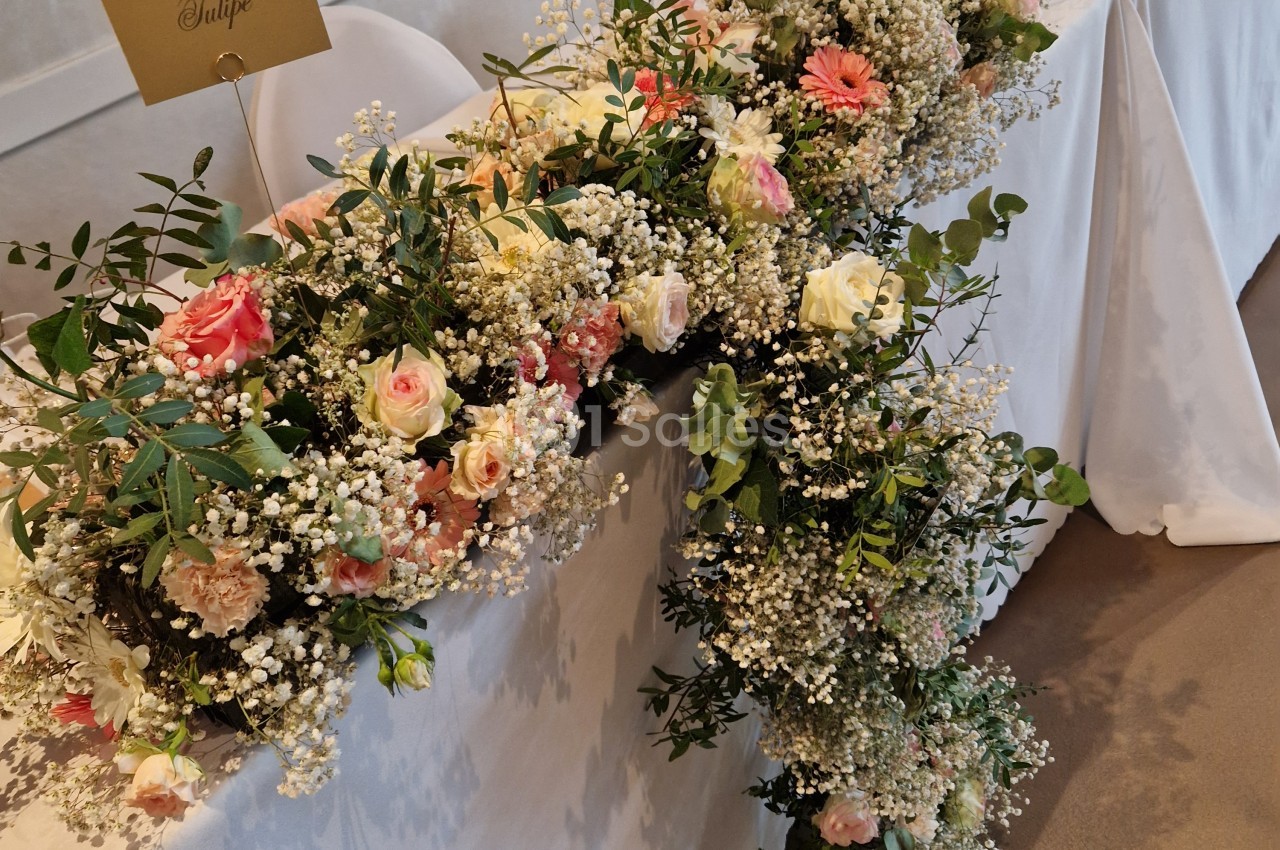 Composition florale en longue guirlande sur une table recouverte de nappe blanche, mêlant roses pêche et crème, gerberas, gypsophile blanc et feuillage vert.