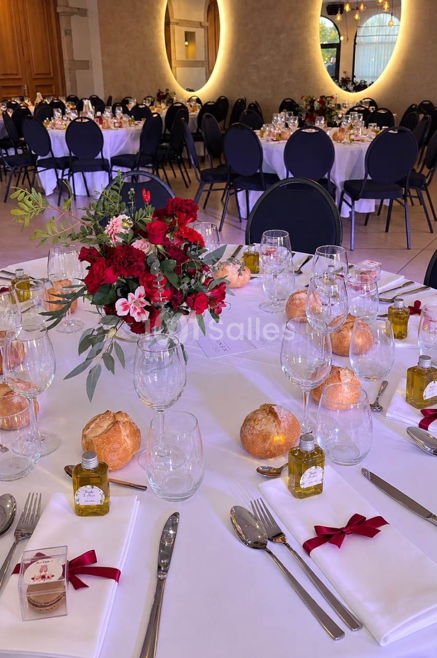 Table de réception dressée de nappes blanches, avec bouquet de roses rouges et fleurs roses, petits pains ronds, bouteilles d'huile d'olive et serviettes nouées de rubans rouges.
