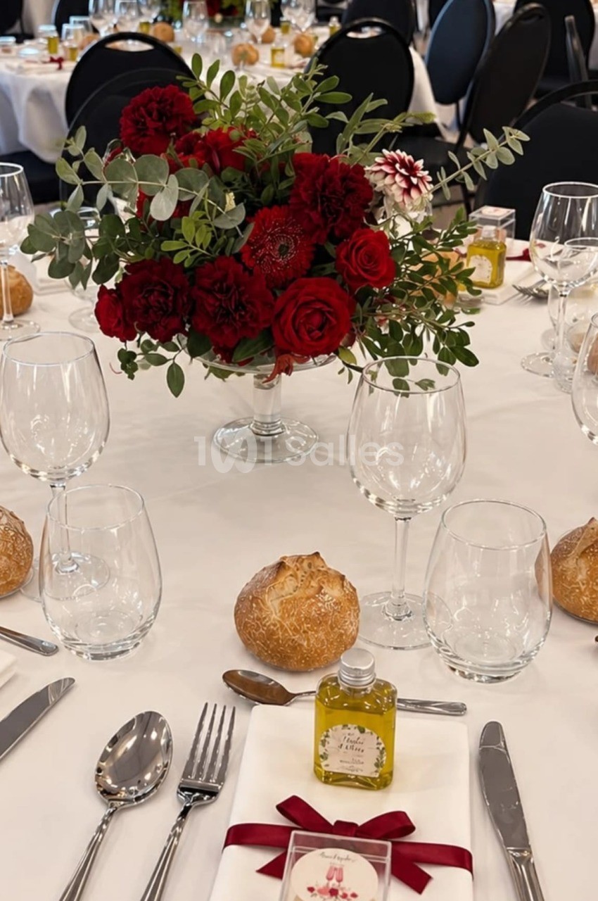 Table de réception dressée de nappes blanches, avec un bouquet de roses et d'oeillets rouges sur pied en verre au centre, verres, couverts et petits pains. Hmm, need to check length: about 150 chars, too long. Shorten. Table ronde dressée de nappe