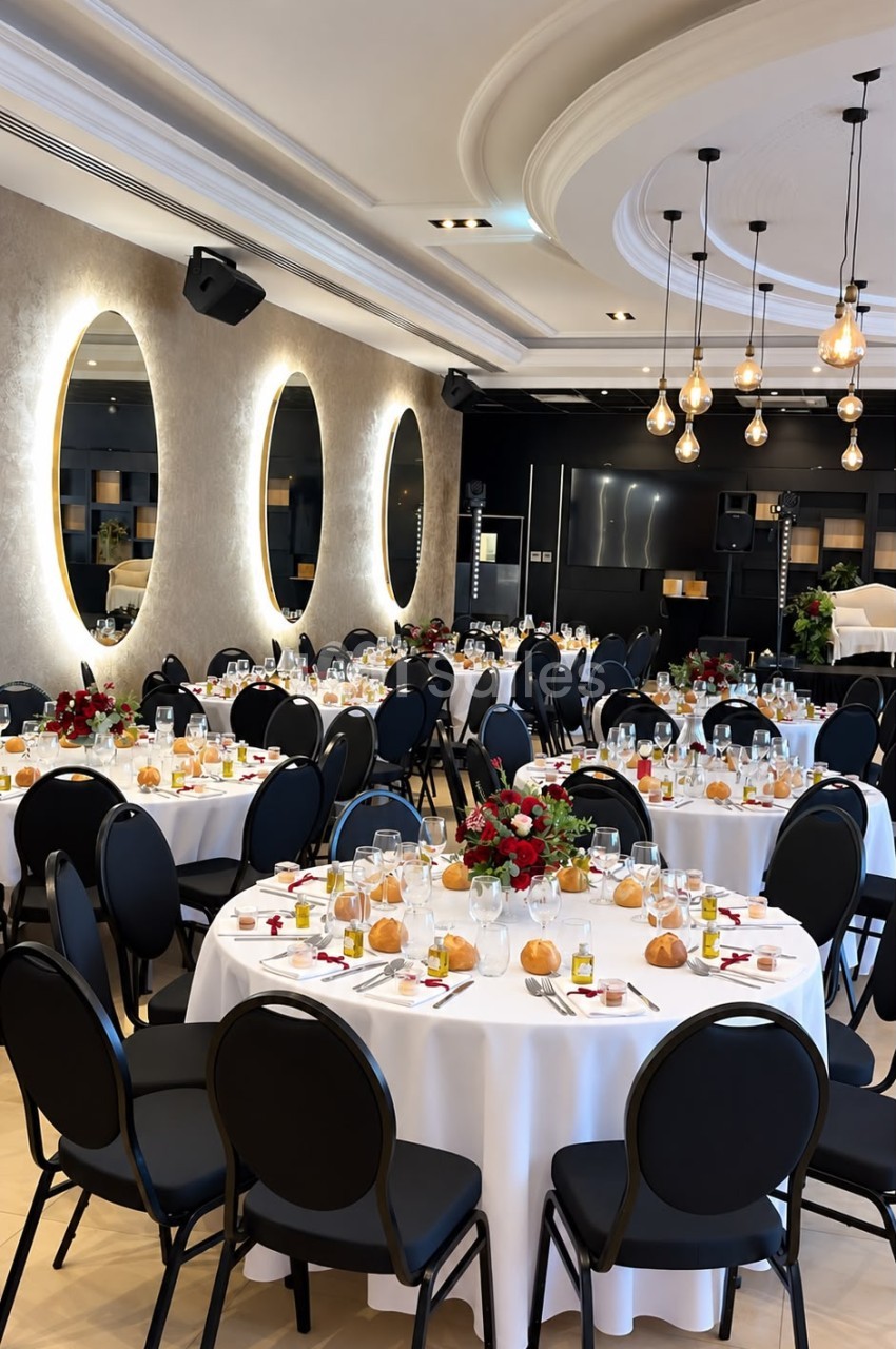 Salle de réception dressée de tables rondes nappées de blanc, chaises noires capitonnées, bouquets de roses rouges et petits pains, sous des suspensions à ampoules apparentes.