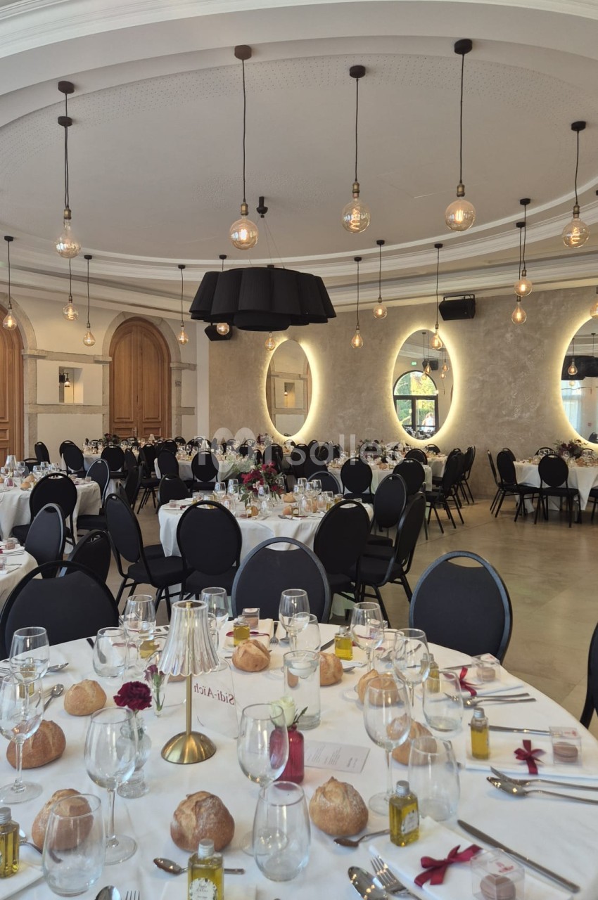 Salle de réception dressée de tables rondes nappées de blanc, avec chaises noires, suspensions à ampoules apparentes et grands miroirs ovales éclairés en fond.