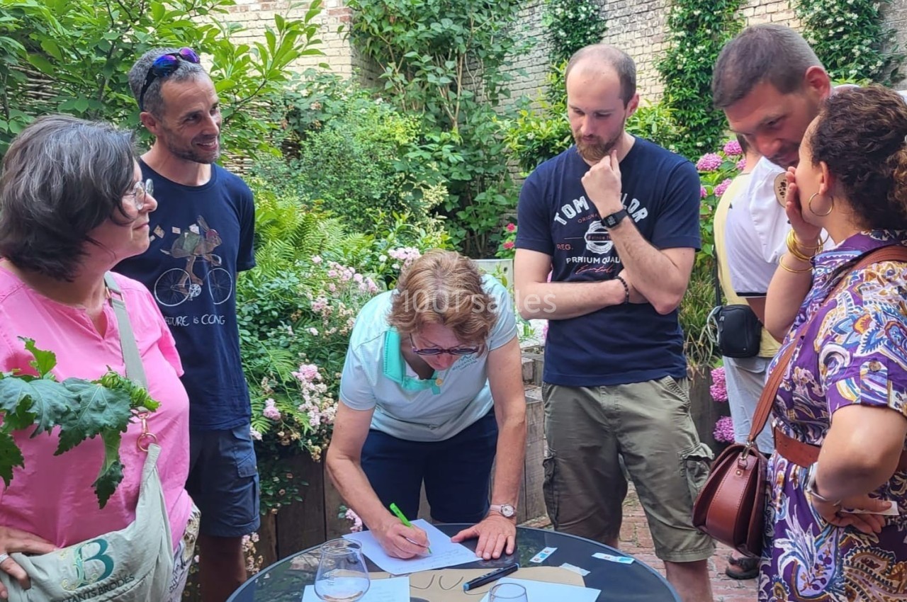 Un groupe de personnes discute autour d'une table en extérieur, dans un jardin fleuri, tandis qu'une personne écrit.