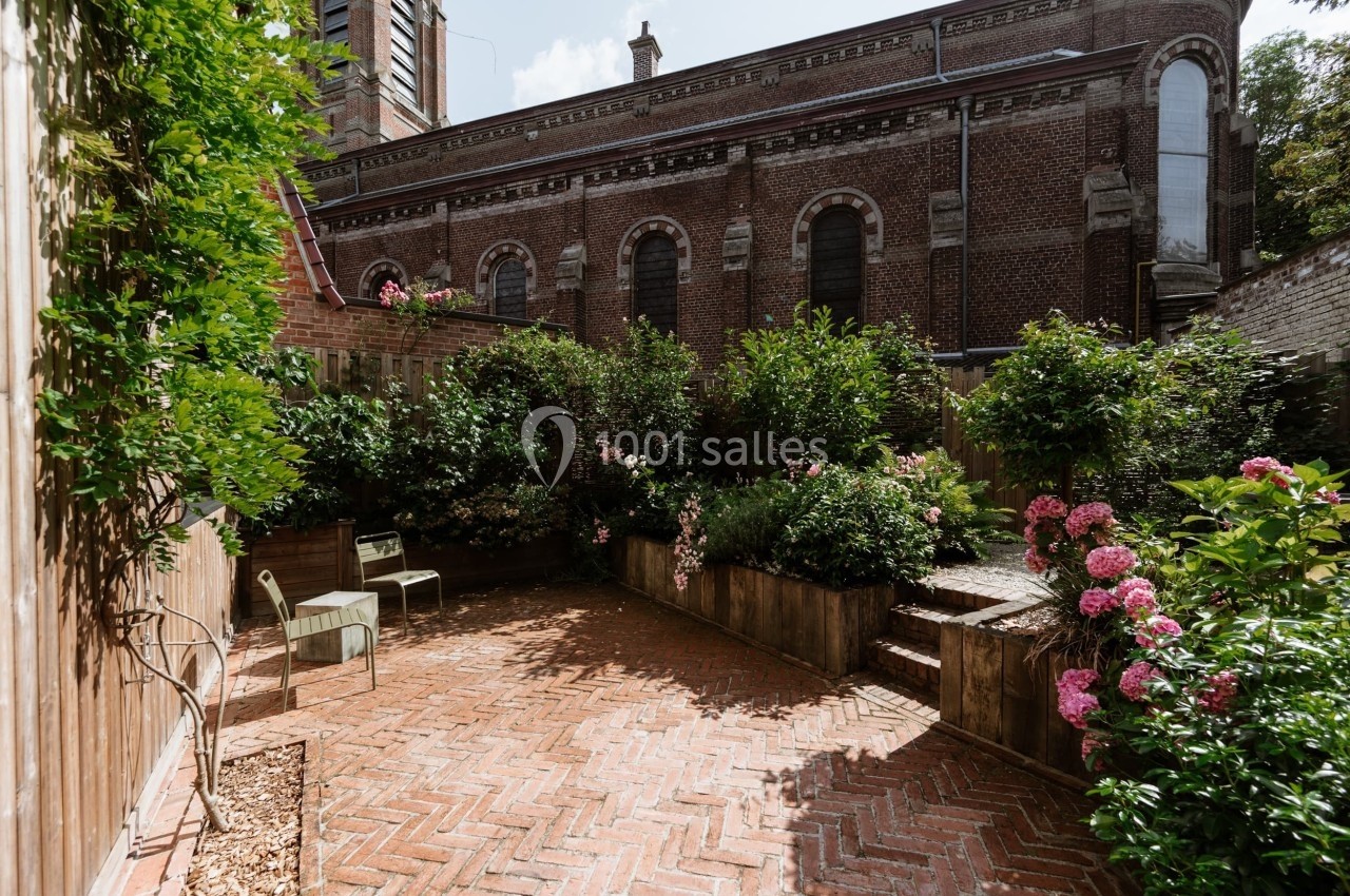 Cour pavée en briques entourée de plantes, avec des chaises et des fleurs roses, adossée à un bâtiment en briques.