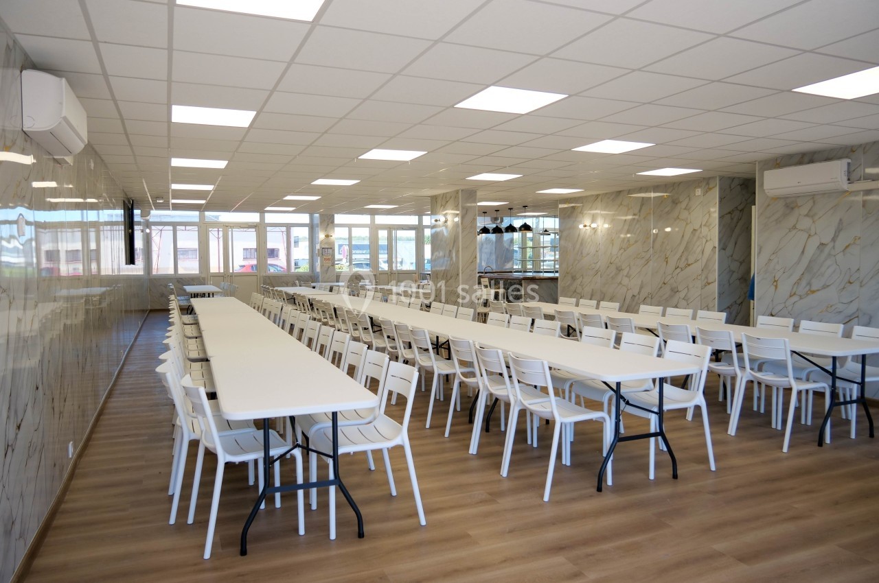 Salle lumineuse avec de longues tables blanches alignées et des chaises, sol en bois et murs à effet marbre.