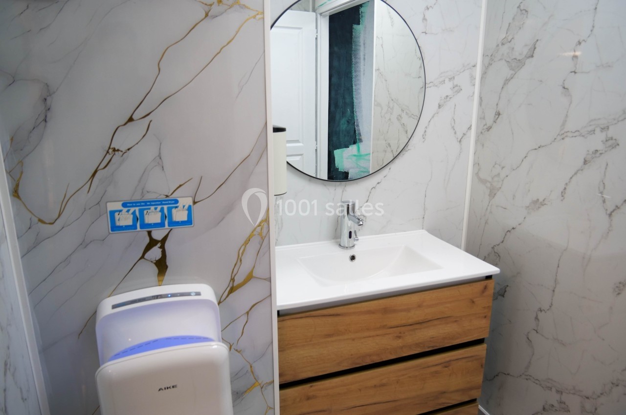 Lavabo avec meuble en bois, miroir rond et mur en carrelage effet marbre, sèche-mains mural à proximité.