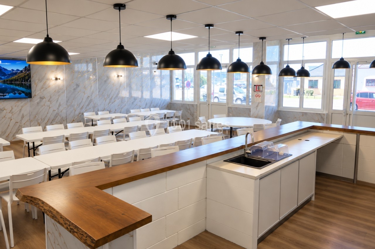 Salle de restauration lumineuse avec tables blanches alignées, comptoir en bois et suspensions noires au plafond.