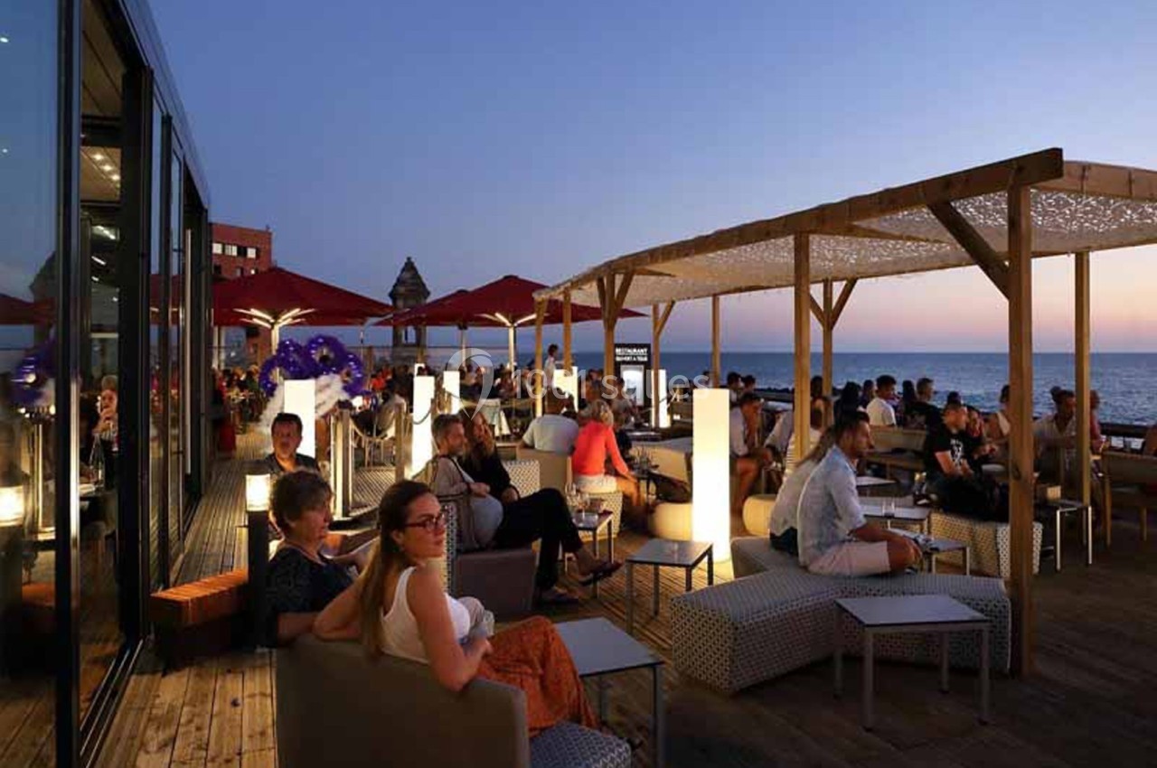 Terrasse en bois au crépuscule, en bord de mer, avec pergola lumineuse, canapés bas et convives assis face à la mer.