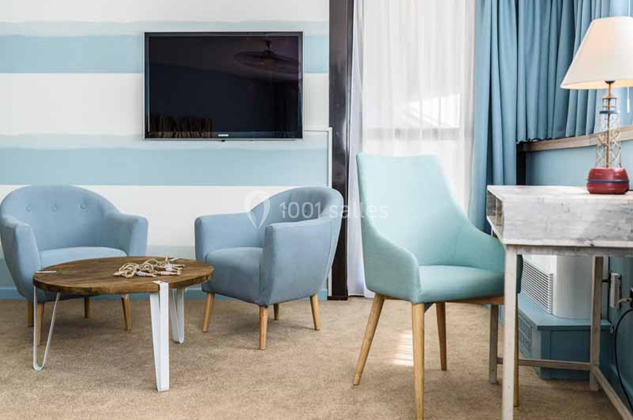 Salon d'hôtel aux murs rayés bleu et blanc, avec deux fauteuils bleus, une table basse en bois, un téléviseur mural et un fauteuil vert d'eau. Let me check length: roughly 150 characters, too long. Need ≤125. Revised: "Coin salon aux murs rayés bleu et