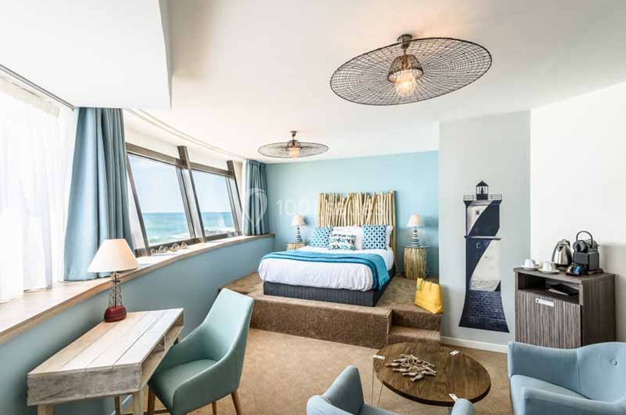 Chambre d'hôtel à dominante bleue avec grand lit sur estrade, fenêtres donnant sur la mer, phare décoratif au mur et table basse en bois.