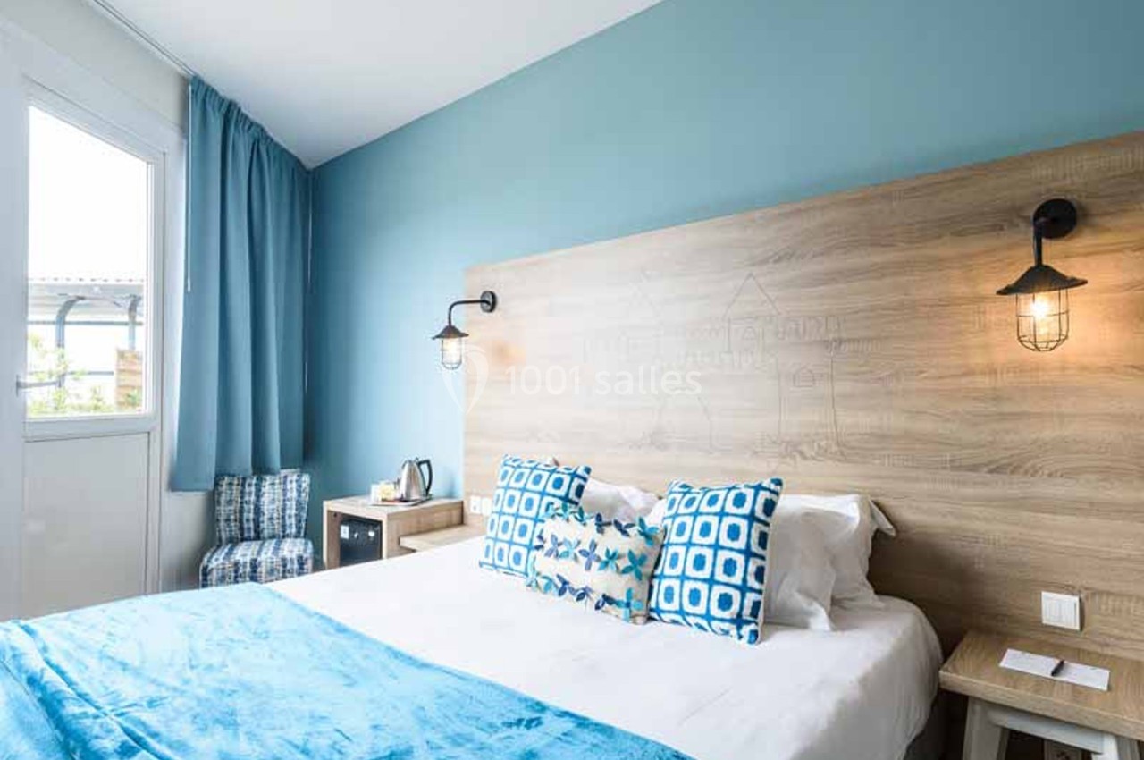 Chambre d'hôtel avec un grand lit recouvert de linge blanc, coussins bleus à motifs et plaid turquoise, tête de lit en bois clair et murs bleu ciel. Let me check: under 125 characters? Count roughly: "Chambre d'hôtel avec un grand lit recouvert de linge