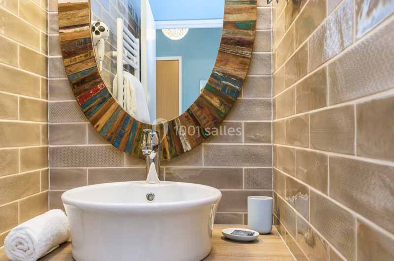 Lavabo à poser blanc sur un plan en bois, miroir rond à cadre de bois multicolore et carrelage beige en relief dans des toilettes.