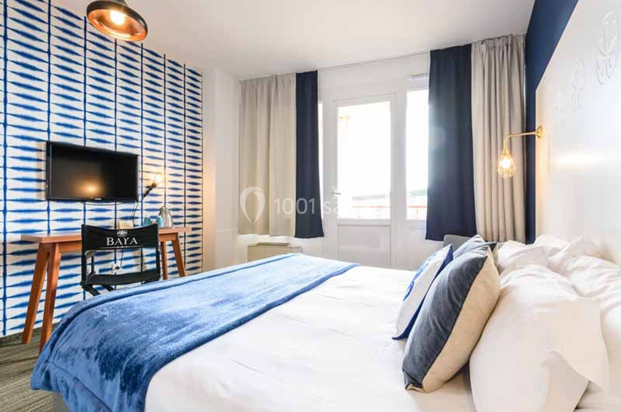 Chambre d'hôtel avec un grand lit recouvert de linge blanc et d'un plaid bleu, un mur à motif rayé bleu et blanc, un bureau en bois et une télévision murale. Let me check length: roughly 165 characters, too long. Need ≤125. Chambre d'hôtel à grand lit