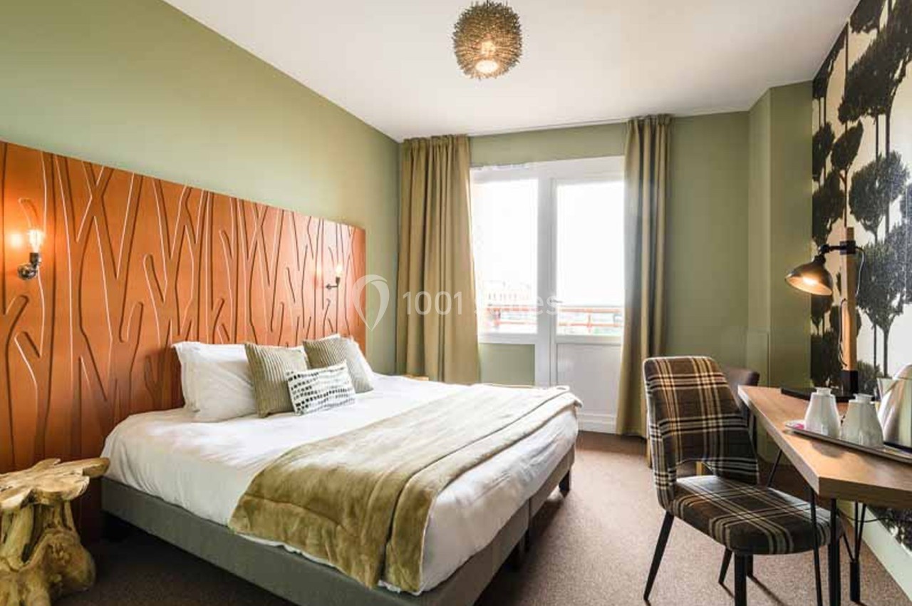 Chambre d'hôtel à lit double avec tête de lit en bois sculpté orange, murs verts, rideaux beige et bureau en bois face à une fenêtre. Let me check length: roughly 125 characters? Count: "Chambre d'hôtel à lit double avec tête de lit en bois sculpté
