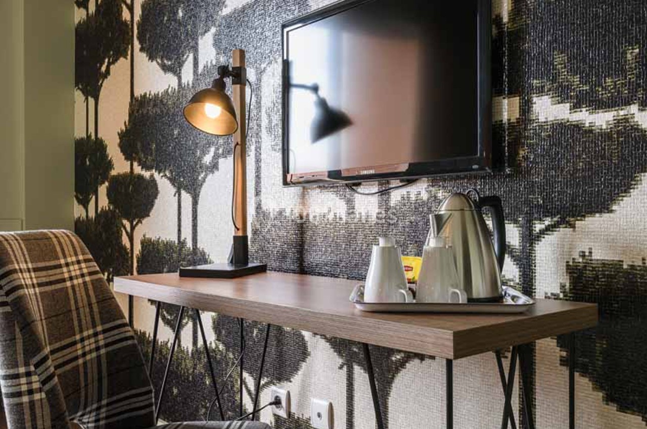 Coin bureau de chambre avec une lampe de table en bois et métal, un téléviseur fixé au mur, une bouilloire et deux tasses sur un plateau, devant un papier peint à motifs d'arbres noirs. Let me check the constraints: French, one sentence, under 125